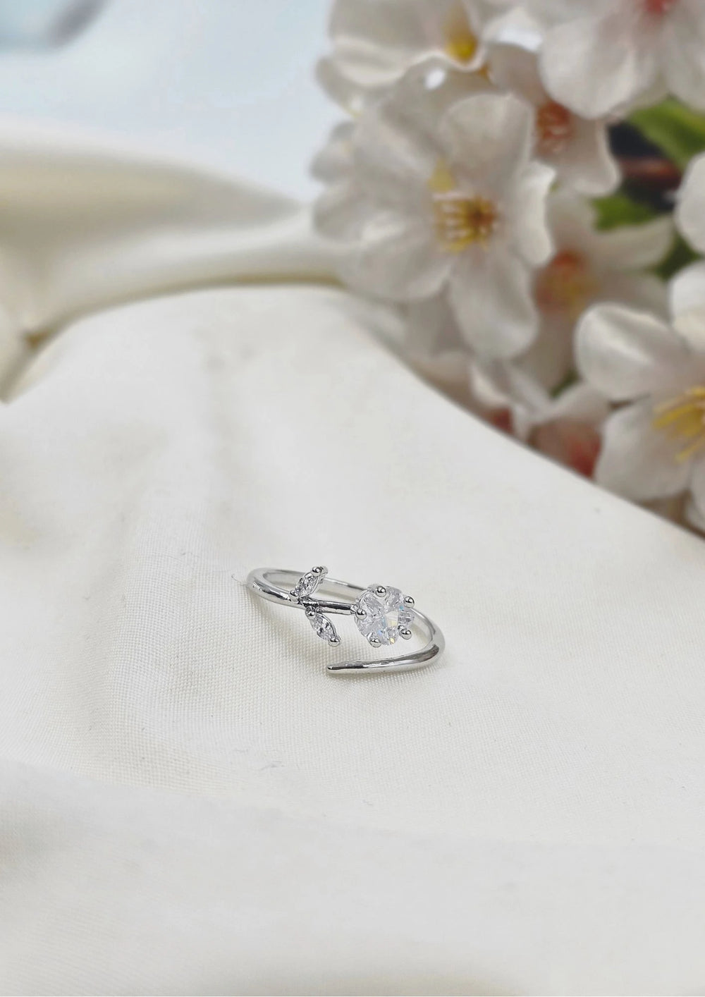 A silver-colored ring with a clear central stone, placed on a white surface with flowers in the background.