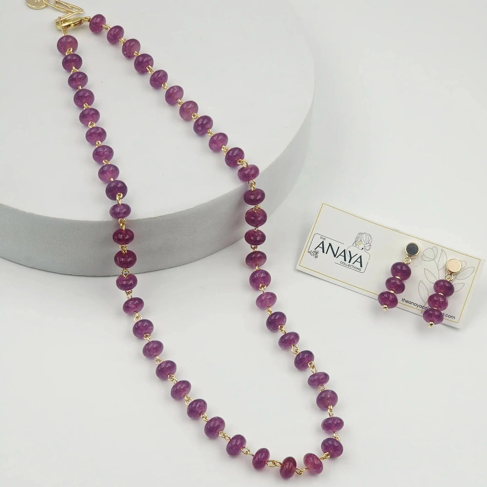 Purple beaded necklace with gold accents on a white surface, accompanied by a matching earring set on a branded card.