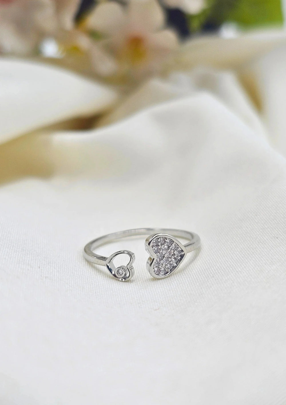 A silver-colored ring with a heart and leaf design, featuring small embedded stones, displayed on a white surface with a floral pattern in the background.