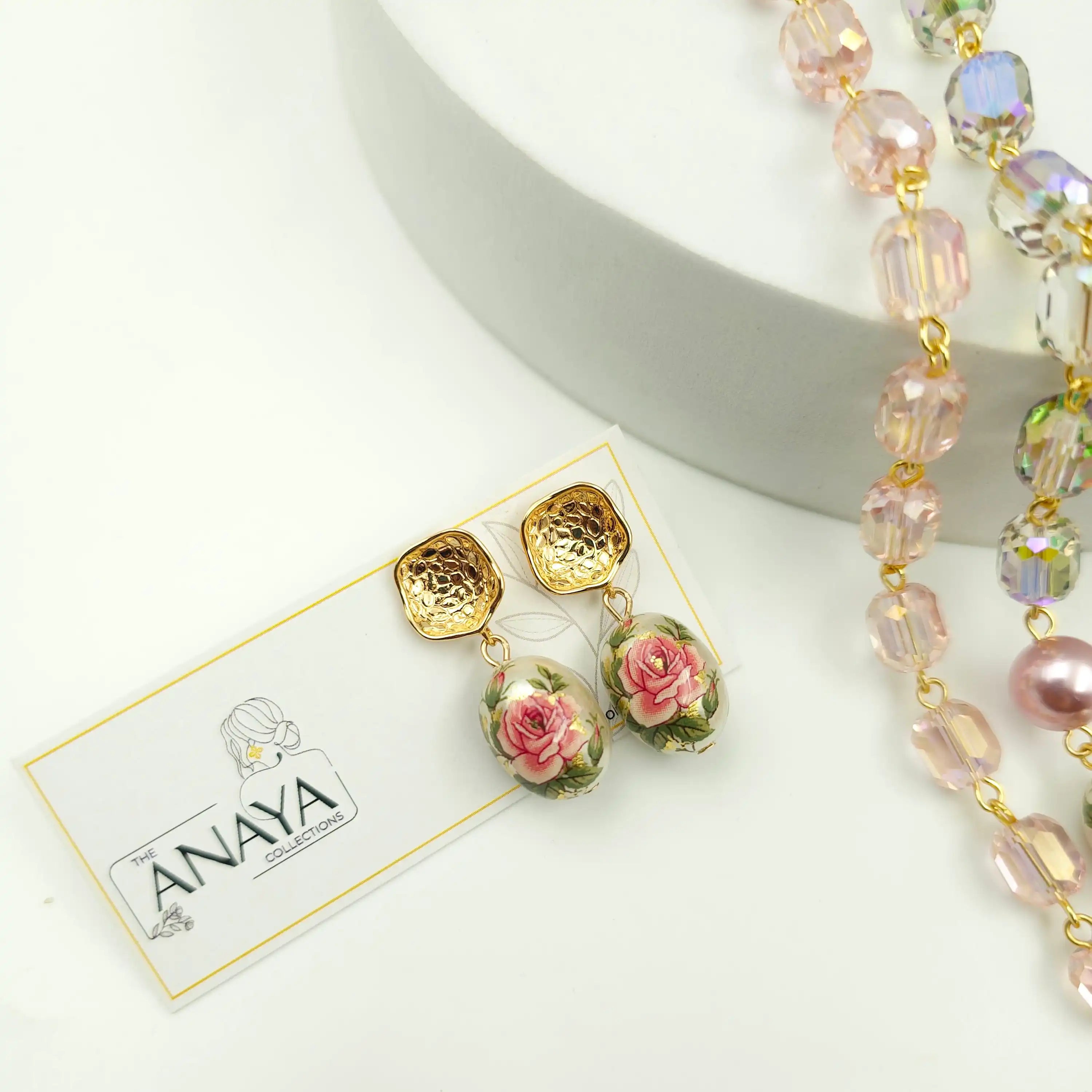 Gold and pink floral earrings on a white card with 'ANAYA' branding, next to a beaded necklace.