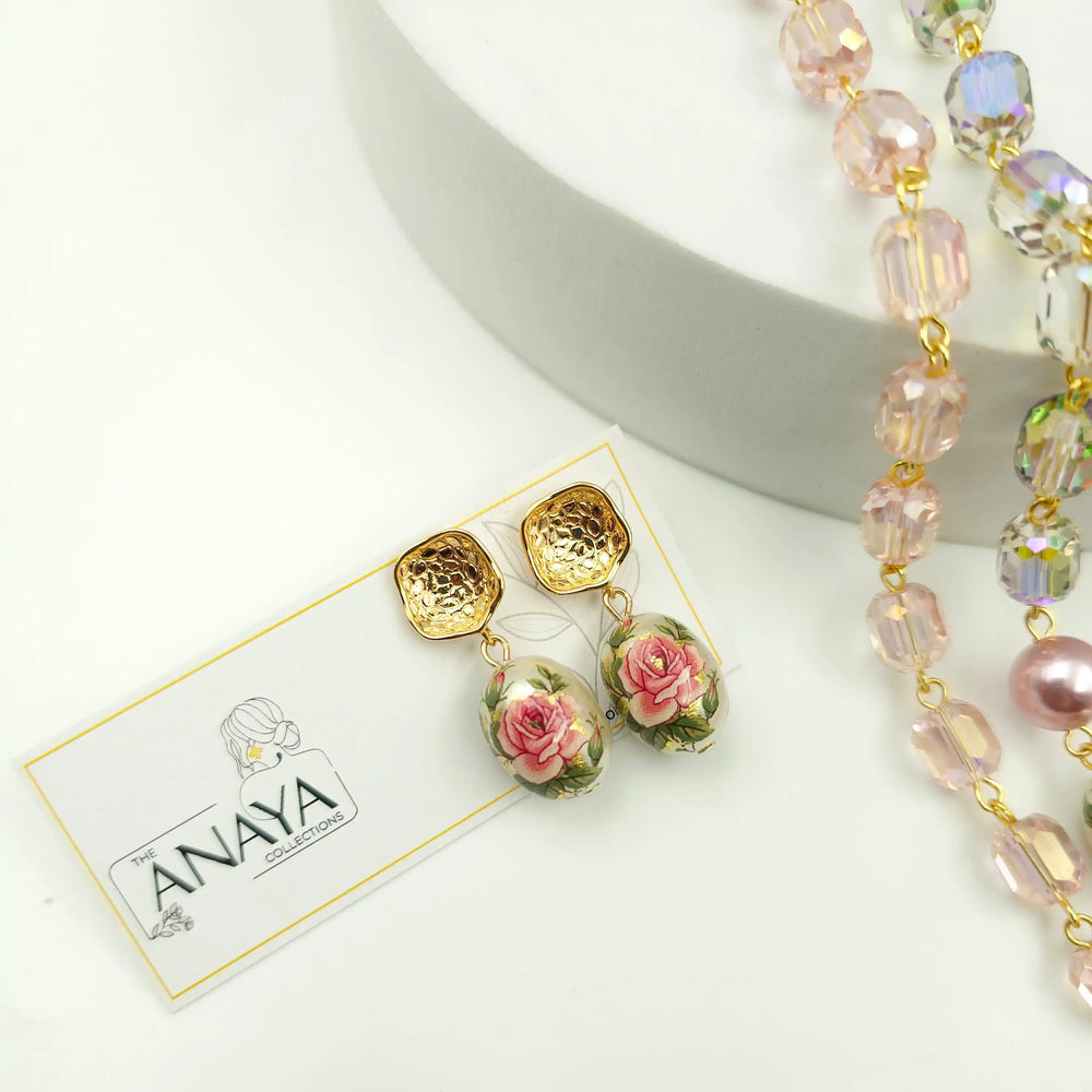 Gold and pink floral earrings on a white card with 'ANAYA' branding, next to a beaded necklace.